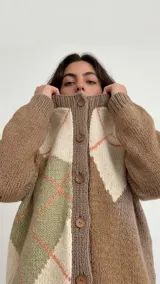 Cárdigan de punto con diseño geométrico de bloques de color en tonos tierra, verde y crema, con líneas finas en color coral. Presenta cierre frontal con botones, cuello redondo y mangas raglán.