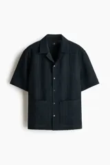 Camisa de manga corta color negro con textura acanalada, cuello tipo resort, cierre frontal con botones y dos bolsillos aplicados al frente.