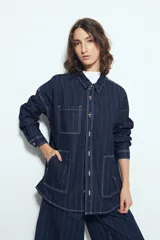 Sobrecamisa de jean color azul oscuro con estampado de rayas verticales blancas. Presenta un corte relajado, cuello clásico, cierre frontal con botones metálicos, mangas largas y dos bolsillos frontales de parche.