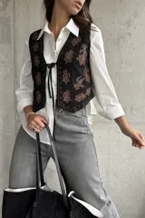 Chaleco negro de gamuza con bordados florales en tonos marrones. Presenta un cierre frontal con lazo y detalles de tachas metálicas en los bordes.