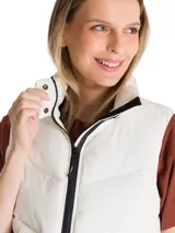 Chaleco puffer corto de color beige, con cuello alto, cierre frontal con cremallera y solapa protectora. Presenta un diseño acolchado, bolsillos laterales con cierre y parche de silicona con logo en la nuca.