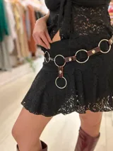 Cinturón de cuero marrón oscuro con acabado brillante, adornado con grandes aros metálicos plateados y un colgante con un aro más pequeño.