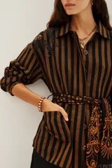 Saco tipo camisa de algodón con rayas verticales marrones y negras. Presenta bordados de cuentas en tonos tierra en el hombro izquierdo y se ajusta a la cintura con un lazo de viscosa estampado en tonos naranjas y negros.