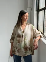Blusa beige de lino con estampado floral en tonos marrones y verdes, escote en V con botón dorado decorativo y mangas largas.