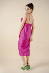 Vestido midi fucsia de satén con escote drapeado, breteles finos y tajo lateral. Presenta una rosa de tela en el bretel izquierdo.