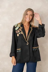 Blazer negro de corte recto con solapas decoradas con ribete dorado trenzado. Presenta bordados artesanales coloridos en el hombro derecho y detalles de flecos con cuentas turquesas en los bolsillos frontales. Cuenta con cierre de un solo botón.