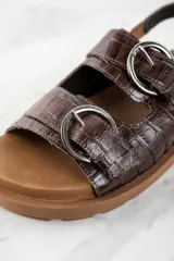 Sandalias bajas color marrón con textura croco, dos tiras anchas sobre el empeine con hebillas metálicas plateadas y tira trasera ajustable con hebilla.