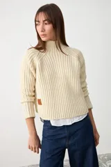 Sweater de punto grueso en color crudo, con cuello alto y diseño de canalé vertical. Presenta mangas largas con corte raglán y un ajuste holgado.