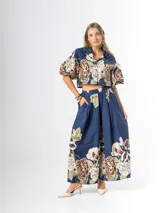 Conjunto de dos piezas confeccionado en viscosa con estampado floral en tonos marrones y verdes sobre base blanca. Incluye una blusa tipo camisa corta con cuello solapa, cierre frontal de botones y mangas abullonadas con diseño globo, y un pantalón tipo culotte de tiro alto, amplio y fluido con pinzas en la cintura.