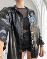 Sobrecamisa negra de cuero sintético con textura croco, corte recto, cuello camisero, cierre frontal con botones a presión plateados, manga larga con puños abotonados, bolsillo de parche en el pecho y largo hasta la cadera.