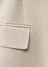 Blazer de lino color beige con diseño cruzado, cuello de pico y hombros estructurados. Incluye un cinturón ajustable con hebilla del mismo tejido y dos bolsillos laterales con solapa.