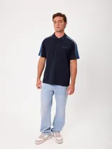 Remera polo gris melange de ajuste holgado, con cuello y cierre frontal de botones. Presenta detalles en azul marino y blanco en los hombros y mangas, y el logo de la marca bordado en el pecho.