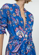 Vestido midi de algodón con estampado floral en tonos azules, rosas y crema. Presenta escote en pico, cierre frontal con botones y un lazo decorativo en la cintura que crea un efecto de nudo. Tiene mangas largas con puños abotonados y un diseño de corte recto.