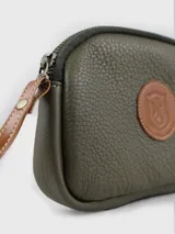 Neceser de cuero color verde oliva, con textura granulada y cierre superior. Presenta un parche circular de cuero marrón con un escudo grabado en el centro. Incluye una correa de muñeca de cuero marrón.
