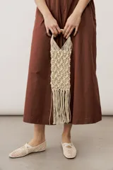 Cartera de mano de macramé color crudo con apliques de caracoles y flecos colgantes en la parte inferior. Cuenta con asas rígidas integradas en el mismo tejido.