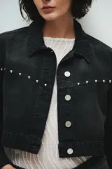 Campera de jean negra, corte crop, con cuello clásico, cierre frontal con botones metálicos y tachas plateadas en hombros y solapas.