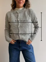Cárdigan de punto gris con efecto jaspeado, diseño de trenzas en las mangas, cuello redondo y cierre frontal con botones. Presenta puños y ruedo acanalados.