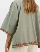 Cardigan tipo kimono de color verde con bordados florales en tono marrón a lo largo de los bordes frontales y en las mangas. Presenta un corte amplio y fluido con mangas tres cuartos.