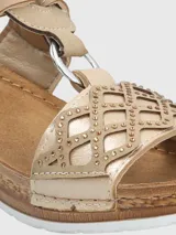 Sandalias de plataforma baja con diseño de tiras cruzadas en color beige y dorado. Presentan una tira superior con cierre de velcro y una tira frontal con detalle de calados geométricos y pequeñas tachas metálicas. La plantilla es acolchada y la suela tiene textura tipo yute.