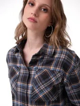 Camisa de corte cropped y diseño holgado, confeccionada en tela de franela con estampado de cuadros en tonos negro, azul y naranja. Presenta cuello camisero, cierre frontal de botones metálicos, mangas largas con puños abotonados y bolsillos en el pecho.