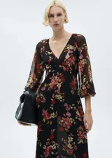 Vestido midi de gasa con estampado floral sobre fondo negro, cuello en pico y mangas largas abullonadas. Presenta un diseño evasé con forro interior y cierre de cremallera lateral.