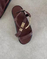 Sandalias planas de color negro con tiras finas y detalle decorativo de tubos metálicos dorados en el empeine. Cuentan con cierre de hebilla ajustable en el tobillo.