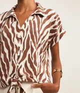Blusa de manga corta confeccionada en viscosa, con cuello camisero, cierre frontal con botones y detalle de lazo para anudar en el bajo. Presenta un estampado híbrido entre follaje y animal print de cebra en tonos marrón y blanco.