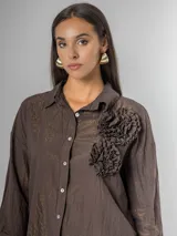 Camisa de manga larga con cuello clásico, confeccionada en tela nacarada con brillo sutil. Presenta apliques de flores tridimensionales fruncidas en el frente, una en color beige claro y dos en color marrón oscuro.