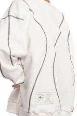 Buzo blanco de manga larga con diseño holgado, cuello redondo y costuras externas visibles en contraste. Cuenta con una etiqueta decorativa en el frente inferior.