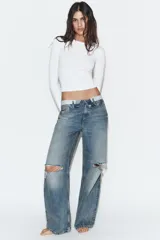 Jeans de tiro medio con diseño de cintura doblada, cinco bolsillos y cierre frontal con cremallera y botón metálico. Presenta un acabado desgastado con roturas en las rodillas y corte recto.