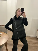 Chaleco negro con cuello solapa y borde de piel sintética, cierre frontal con cremallera y bolsillos laterales con cremallera.