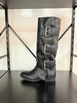 Bota de caña alta confeccionada en ecocuero negro, con diseño de tres hebillas metálicas decorativas en el lateral externo y taco bajo.