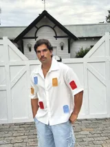 Camisa blanca de manga corta con parches de tela de colores (azul, rojo, amarillo) distribuidos irregularmente.