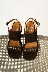 Sandalias de gamuza color marrón con taco cuadrado de madera. Presentan dos tiras anchas, una superior lisa y una inferior decorada con ojales metálicos plateados. Cuentan con cierre de pulsera con hebilla ajustable en el tobillo.