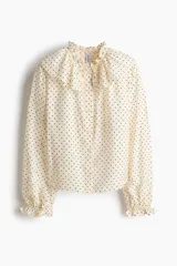 Blusa de viscosa color beige con estampado de lunares negros. Presenta cuello con volados, cordón fino para ajustar, botonadura frontal y mangas largas con puños elásticos fruncidos.