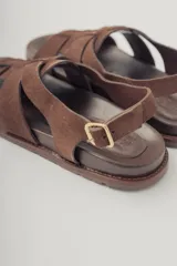 Sandalias planas de gamuza color marrón, con diseño de tiras cruzadas en el empeine y cierre con hebilla metálica dorada en el tobillo. Cuenta con plantilla de cuero y suela de goma con textura.