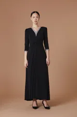 Vestido largo negro de corte elegante con escote en V y mangas tres cuartos. Presenta detalles de pedrería brillante en el borde del escote y en los puños, con un diseño drapeado en la cintura que aporta movimiento y realza la silueta.