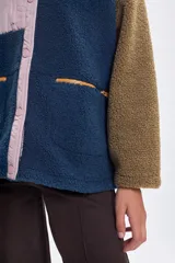 Campera de sherpa con diseño color block, combinando cuerpo azul, mangas marrones y detalles en rosa pálido. Presenta cierre frontal con botones a presión, bolsillo superior con cierre y bolsillos laterales con cremallera.