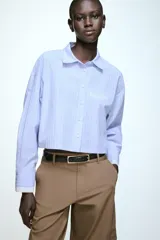 Camisa corta de corte recto con estampado de rayas verticales en tonos azul y blanco. Presenta cuello solapa, cierre frontal con botones, bolsillo de vivo en el pecho y puños anchos en color blanco a contraste.