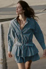 Vestido corto de manga larga con cuello solapa y cierre frontal de botones a presión. Presenta un diseño a cuadros en tonos azul y marrón, cintura ajustable con cinturón de hebilla y falda con volantes en el bajo.