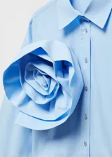 Camisa crop de color celeste con cuello camisero, manga larga con puños abotonados y cierre frontal de botones. Destaca por una maxi flor decorativa aplicada en el pecho.