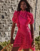 Vestido corto fucsia de encaje, con cuello alto, mangas cortas con volantes asimétricos y falda con diseño cruzado.