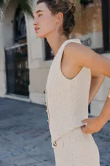 Chaleco de tejido texturado en tonos claros (blanco/beige/gris) con escote pico y cierre frontal de cuatro botones dorados. Presenta un bajo asimétrico.