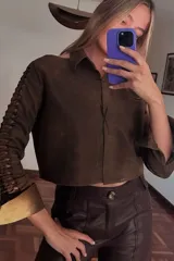 Camisa corta marrón de cuero ovino desgastado, con cuello clásico, cierre frontal con botones, mangas largas con cutouts y puños anchos dorados.