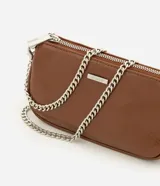 Cartera tipo baguette pequeña Satinato, confeccionada en PU color marfil, de formato alargado y estructura maleable. Cuenta con cierre superior de cremallera, placa frontal aplicada con el logo de la marca y asa de cadena doble metálica en tono plateado que permite llevarla al hombro o cruzada.