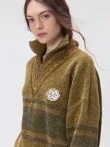 Pullover de textura tipo terciopelo con cuello alto y cierre frontal parcial. Presenta un diseño a rayas horizontales en tonos grises y negros, con detalles en rojo y blanco en el cuello, puños y ruedo. Incluye un aplique bordado con la letra M en el pecho y puños acanalados.