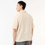 Camisa de manga corta color beige con cuello tipo campamento, cierre frontal de botones y diseño de textura en punto fantasía.