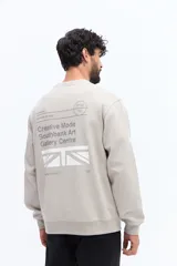 Buzo de algodón y poliéster color verde musgo, con cuello redondo y puños acanalados. Presenta un parche estampado rectangular con texto y un logo circular en el pecho.