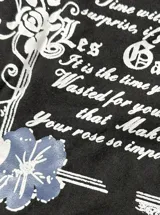 Remera negra con diseño ajustado de manga corta y escote off-shoulder, que incluye un detalle superpuesto simulando una bikini blanca por debajo. Posee estampado gráfico al frente con motivos florales y texto.