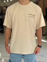 Remera oversize color beige, con mangas cortas y cuello redondo. Presenta un diseño estampado con texto en la parte trasera y lateral, de corte holgado y estilo urbano.